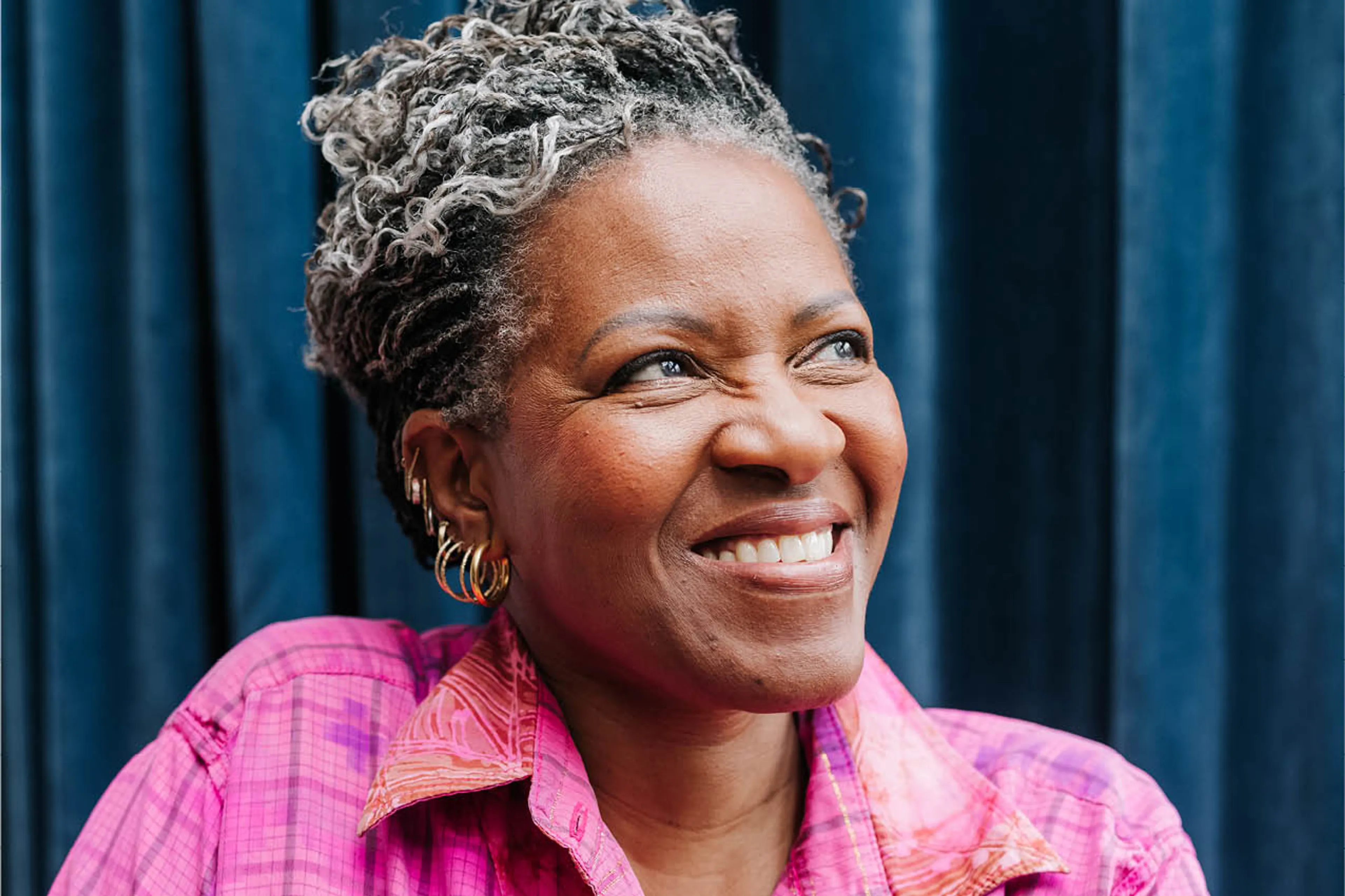 Mpho Tutu lacht breeduit en kijk weg van de camera. Haar felroze blouse steekt contrasterend af tegen de donkerblauwe gordijnen
